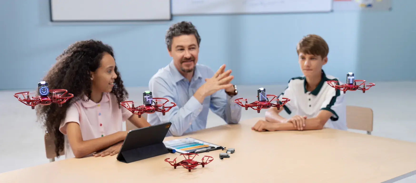 A teacher discusses drones with two students in a classroom to encourage STEM education and coding skills.
