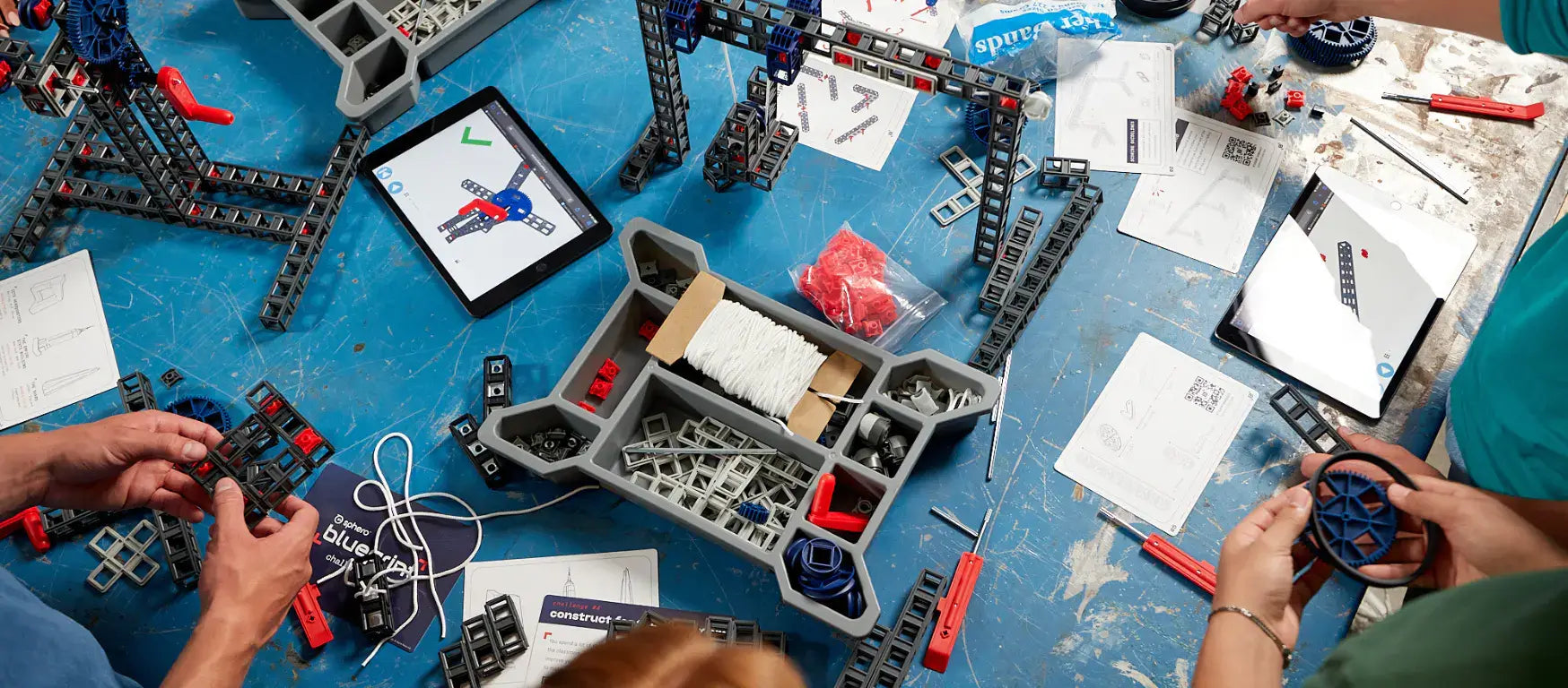 Hands assembling Sphero Blueprint mechanical parts on a blue tabletop with instructional materials and tools.