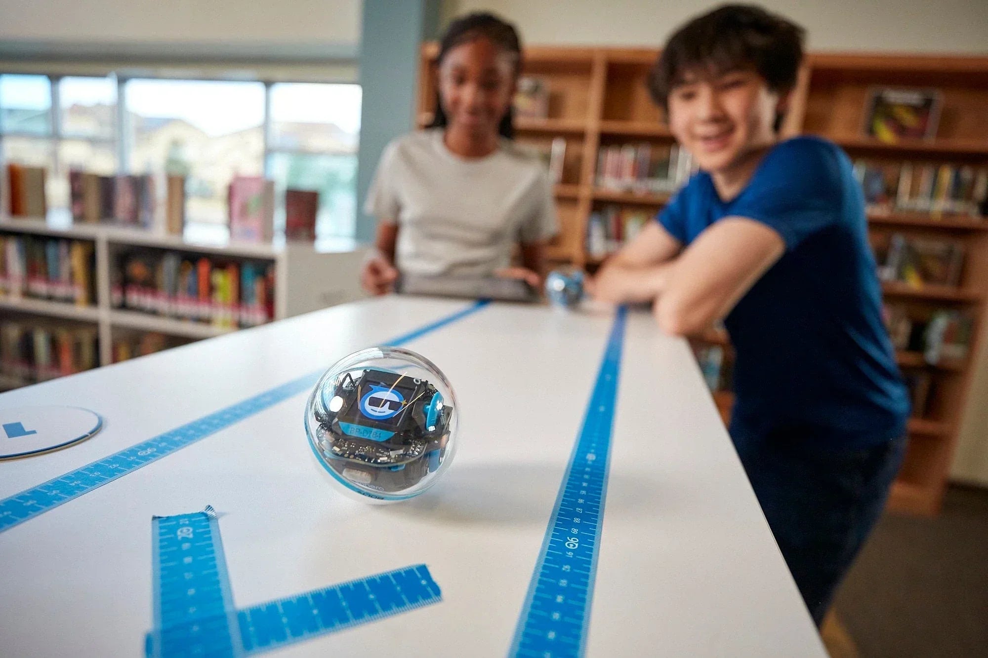 Students engaging with Sphero robotics on a desk in a library, promoting STEM education and interactive learning.