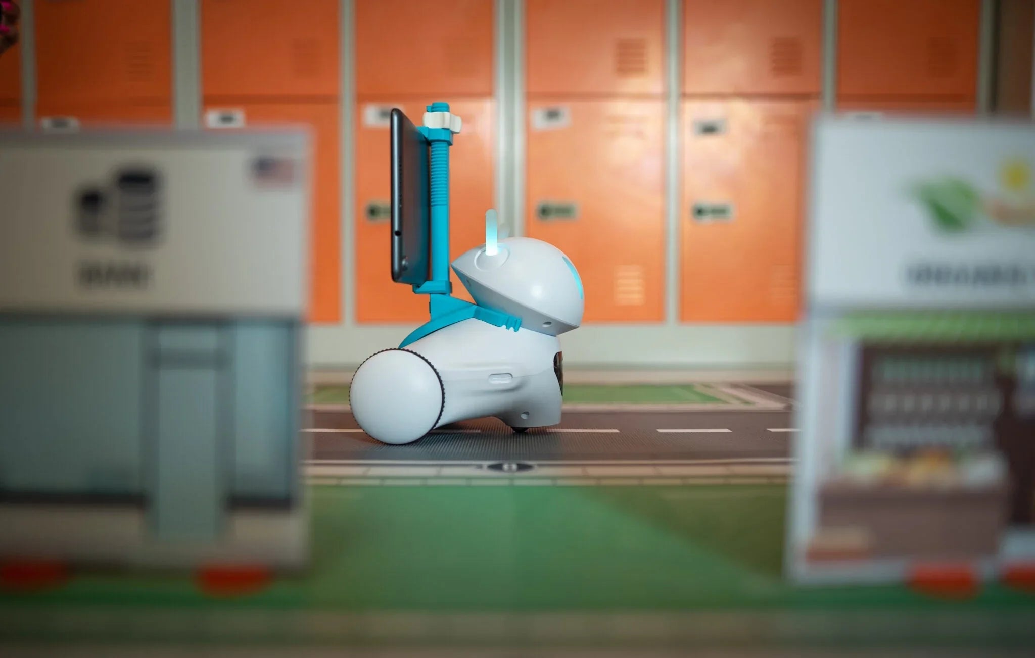 A small robotic device with a monitor, positioned on a toy road amidst miniature buildings and lockers.
