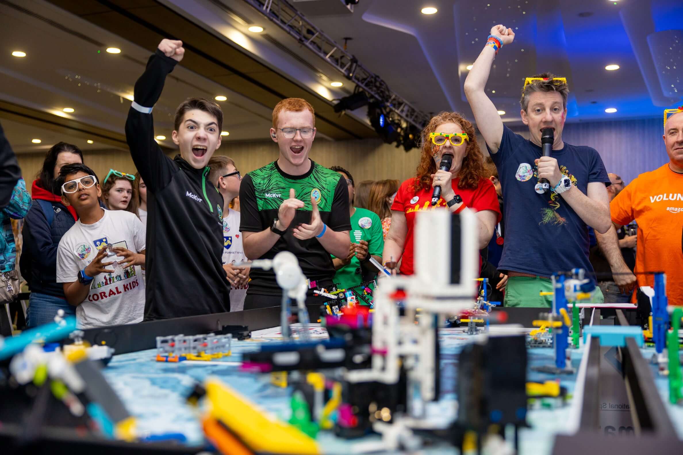 Enthusiastic participants celebrate at a robotics competition with colorful shirts and accessories, amid vibrant LEGO creations.