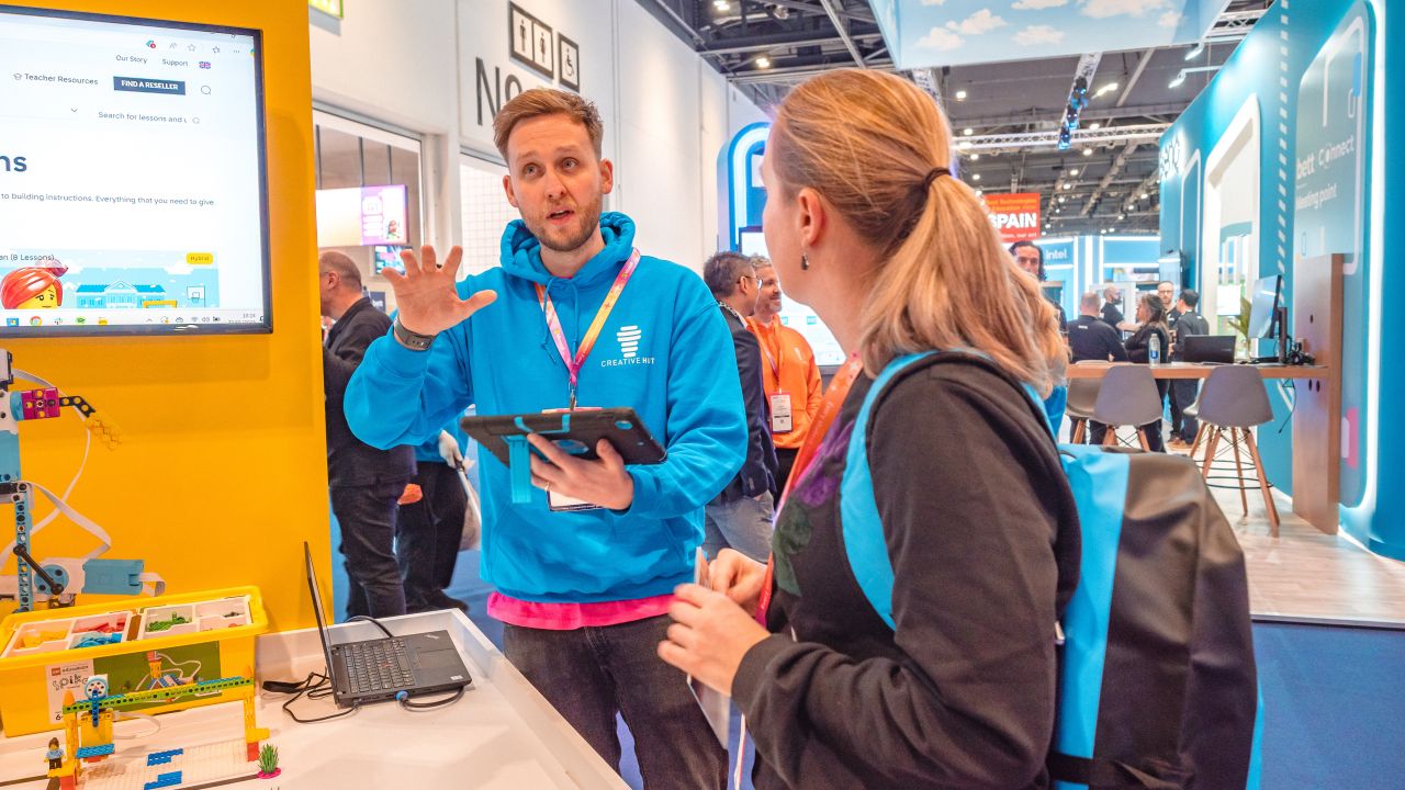 A presenter in a blue hoodie explains a product demo to an attendee at a tech event, with a laptop and models in view.