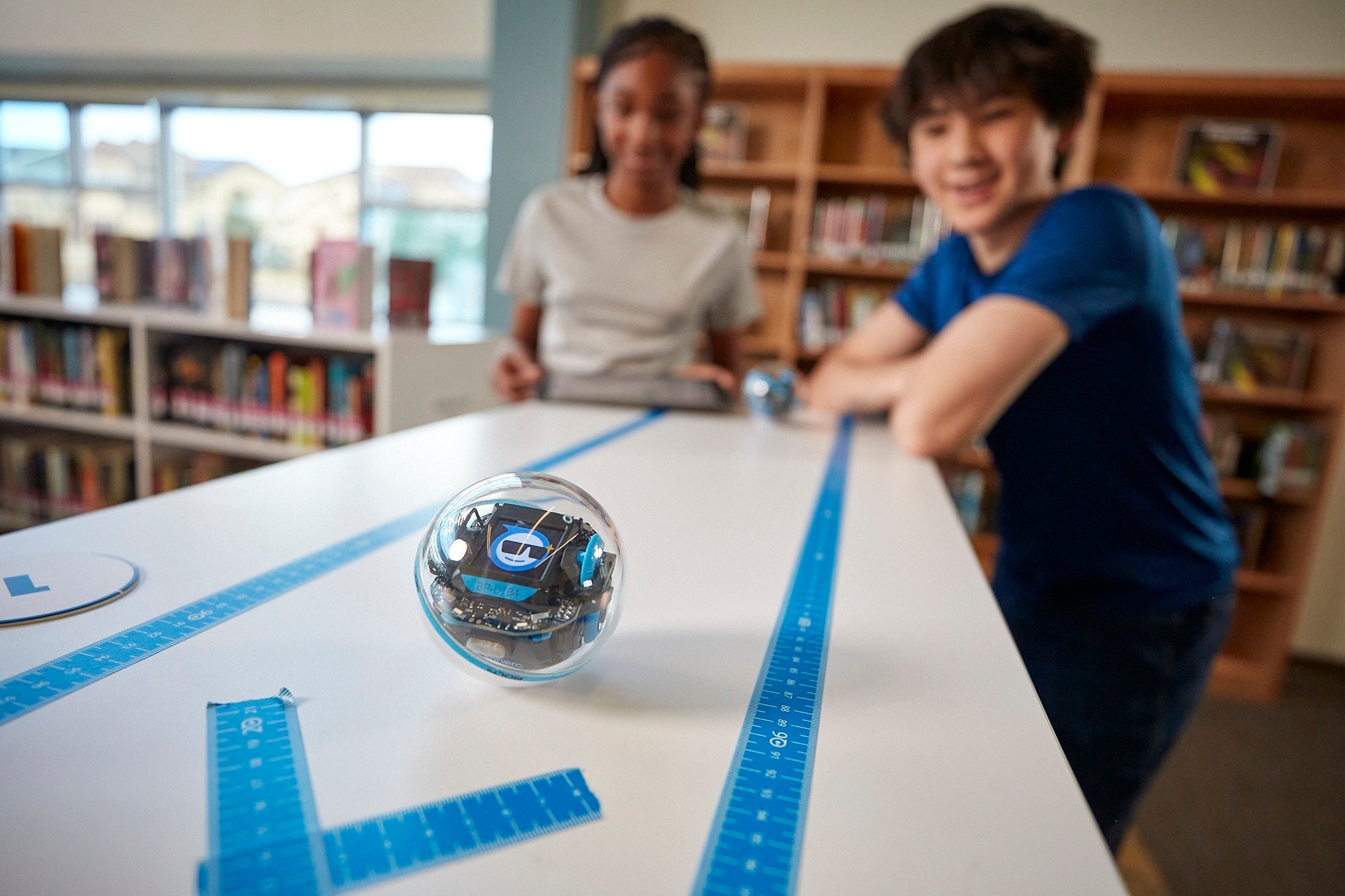 Two children engaging with a robotic ball on a table marked with rulers in a library.