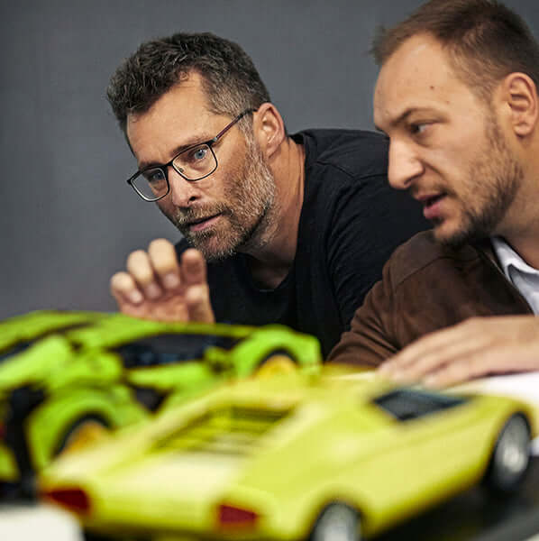 Two men discussing scale models of green sports cars on a table, focused on design details.