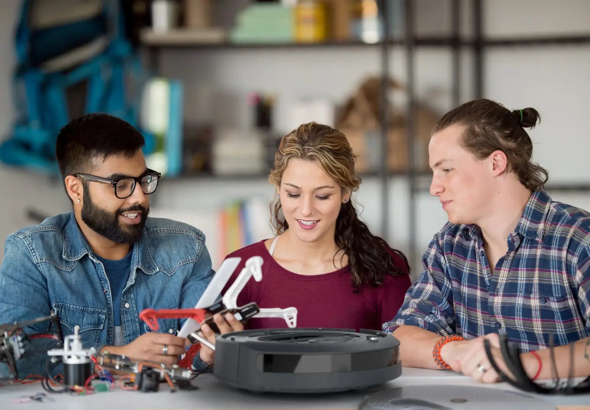 Three young adults collaborating around robotics and technology, discussing drone components and a robot vacuum.
