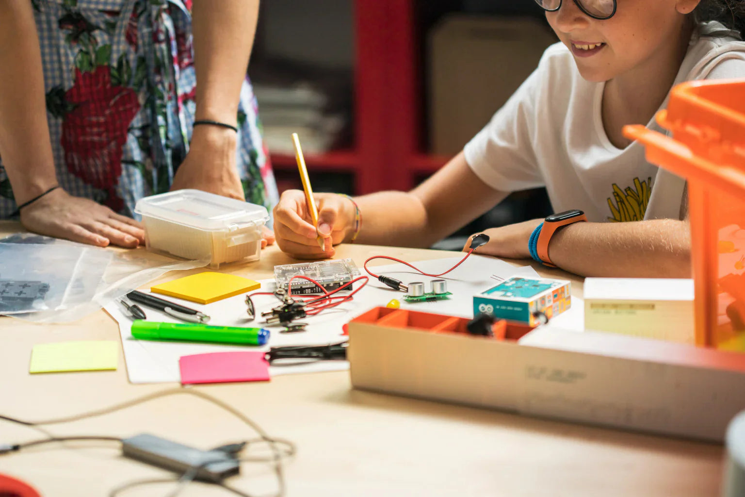 A child engaging in a hands-on electronics project with various components, tools, and stationery on a table.