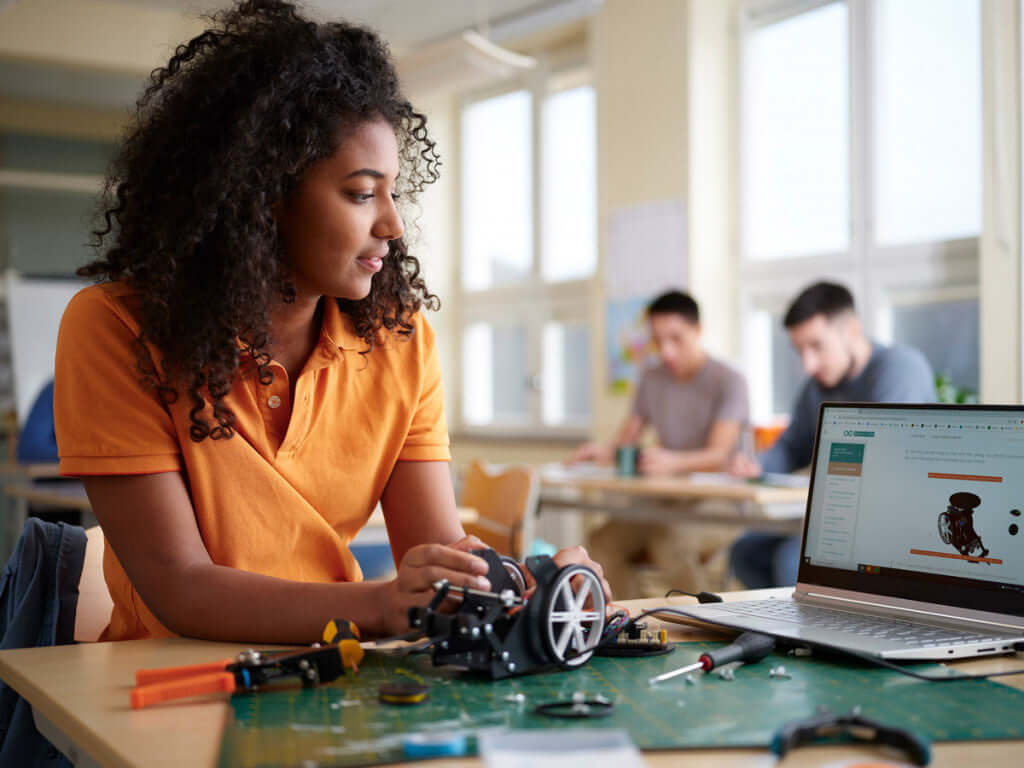 Young woman working on a mechanical project with tools and a laptop in an educational setting.