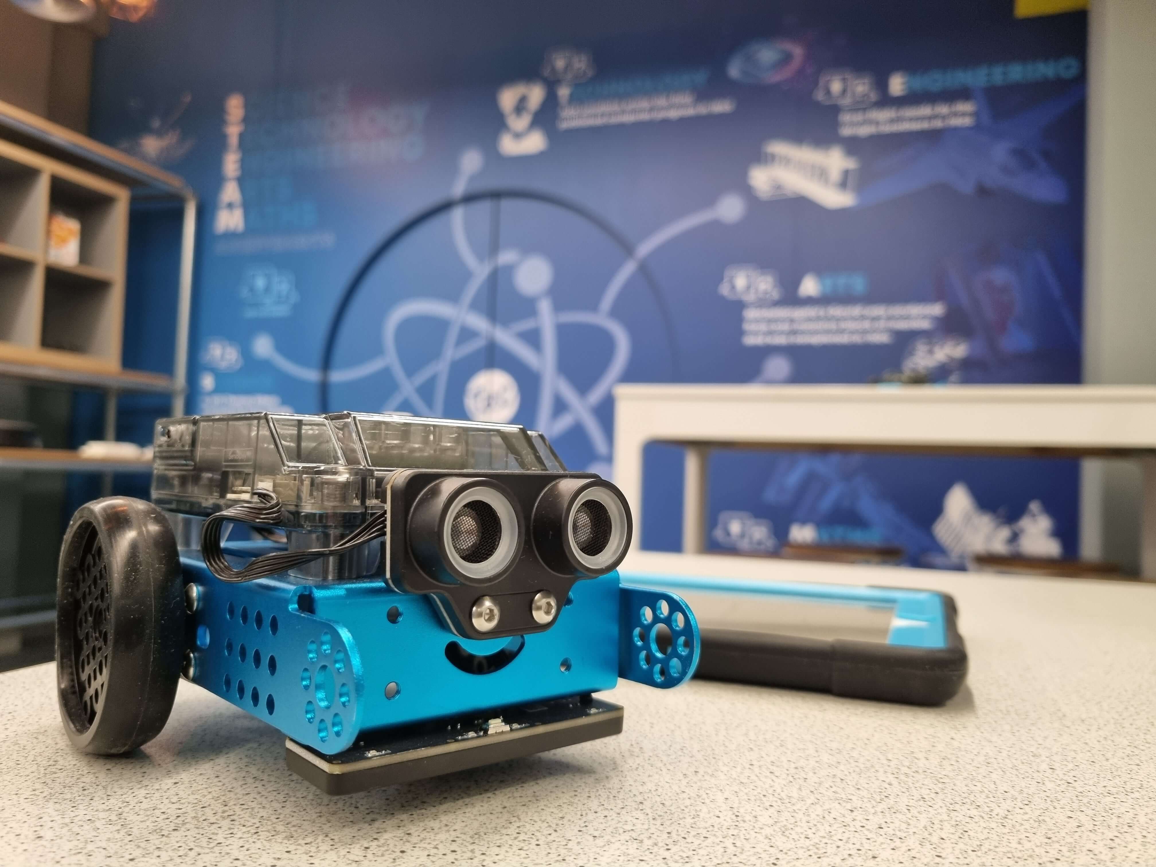 A small blue robot with wheels and eyes, positioned on a table with a science-themed backdrop.