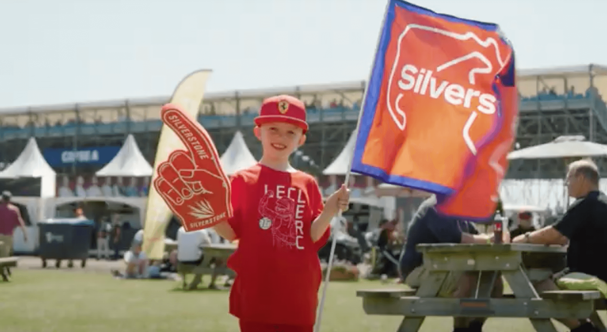 Young fan in a red shirt waving a flag and foam finger at an outdoor event, surrounded by spectators.