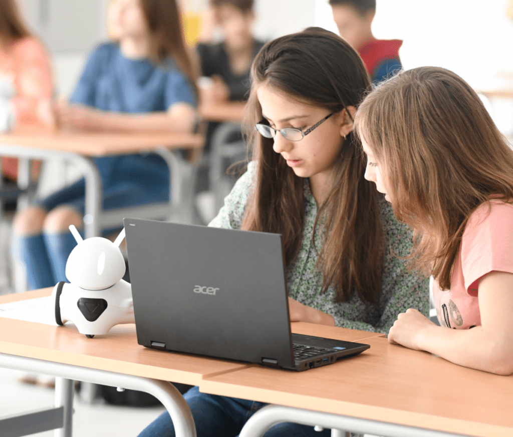 Two students collaborating on a laptop in a classroom setting, with a robotic assistant on the desk.