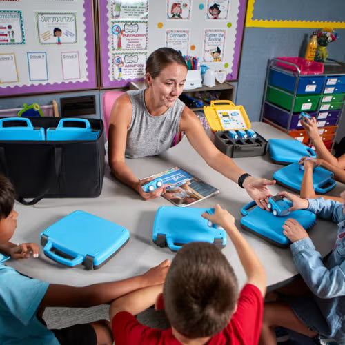 Teacher engages with students at a table, sharing blue learning kits in a classroom setting.