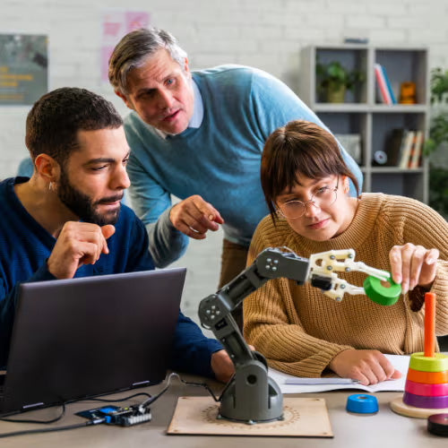 Three people working together on a robotics project with a robotic arm, laptop, and colorful toys in a bright classroom.