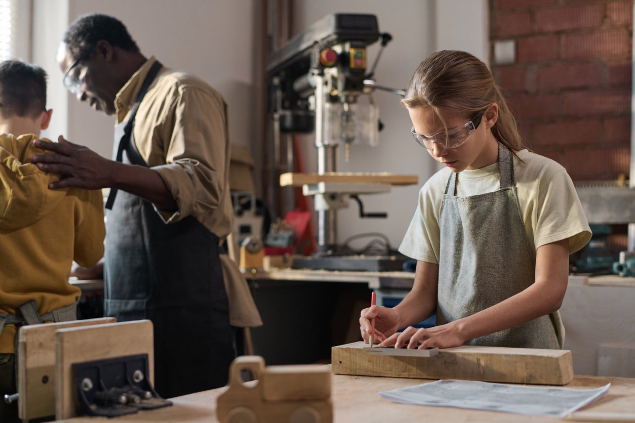 A young girl in safety glasses works on a wooden project in a workshop, guided by an adult.