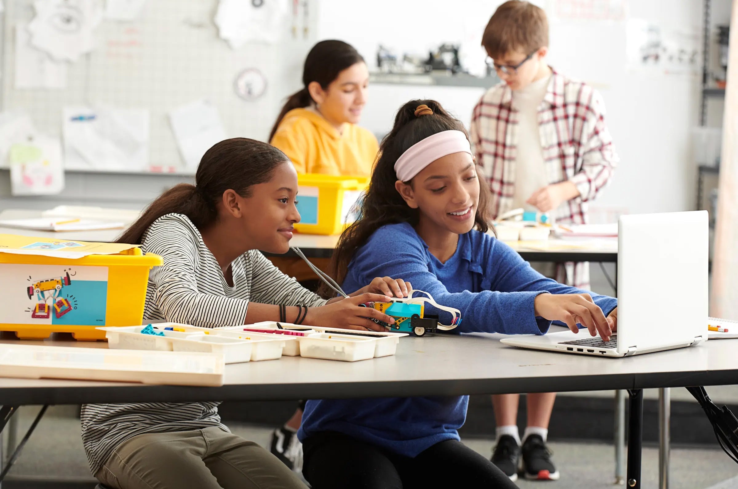 Two girls collaborating on a robotics project at a classroom table, surrounded by classmates and learning materials.