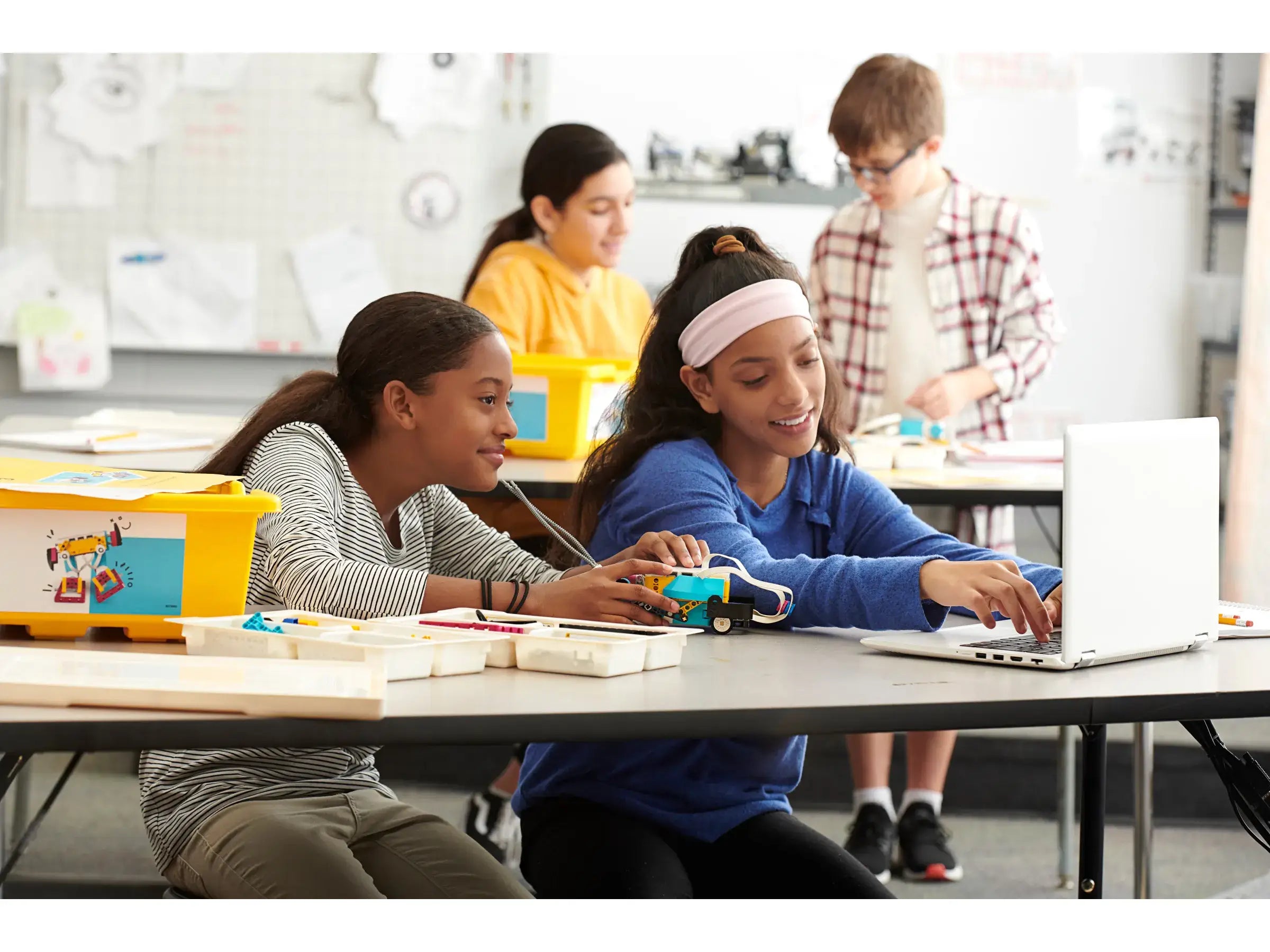 Two girls actively collaborating on a robotics project at a classroom table, with laptops and building materials.