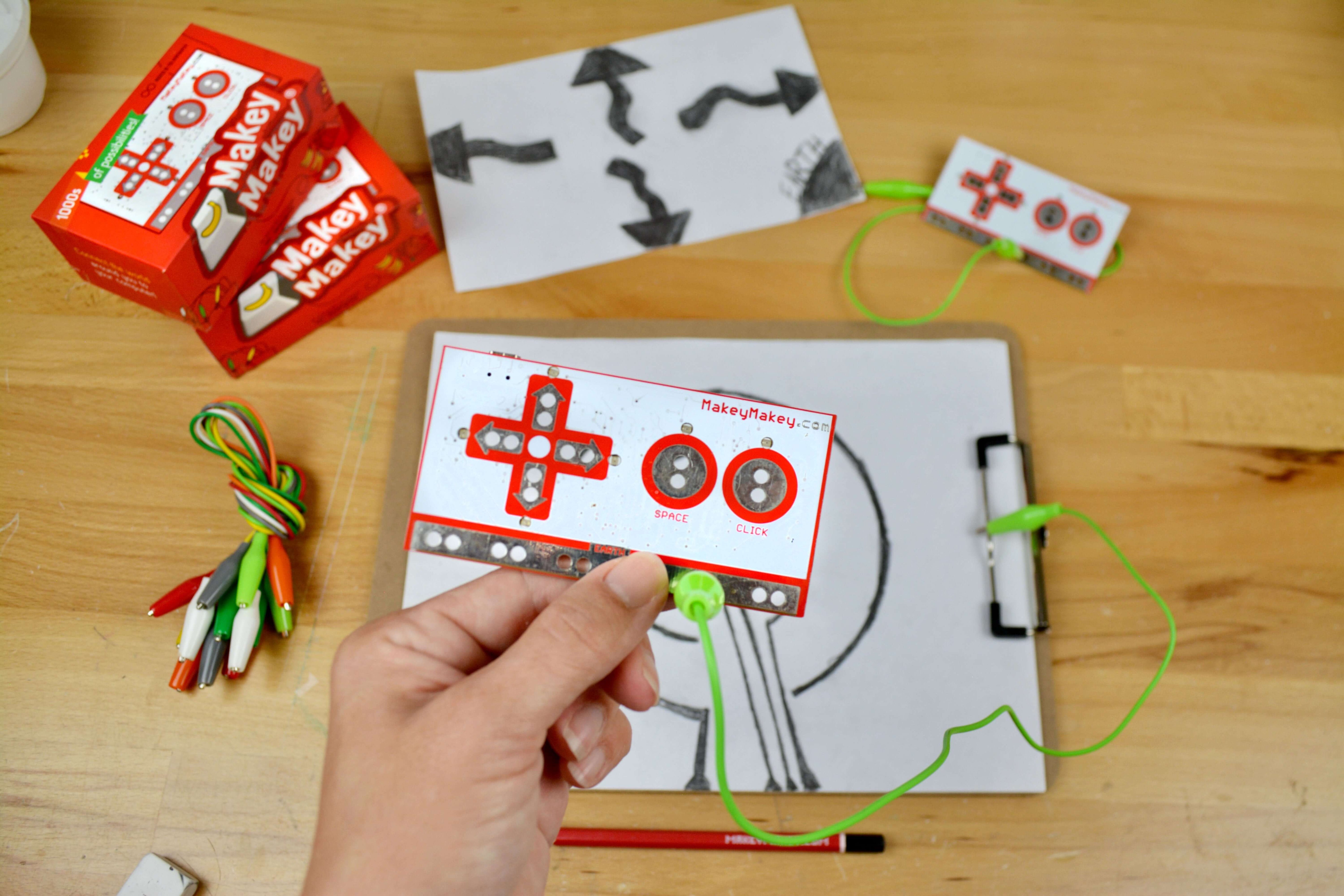 Person holding Makey Makey® circuit board with drawings and tools on a table, demonstrating creative invention possibilities.