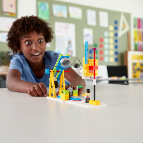 Child excitedly building with colorful LEGO construction set on a table in a bright classroom.