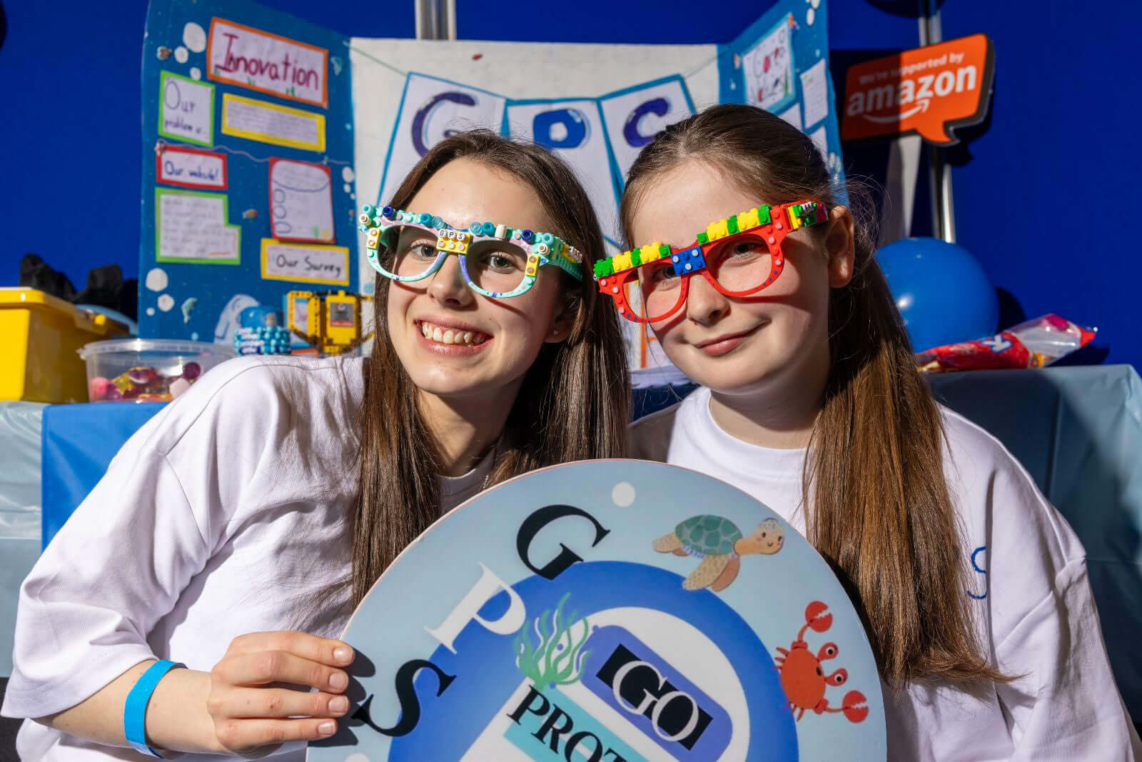 Two girls wearing colorful glasses pose with a project display about marine life and innovation at an exhibition.