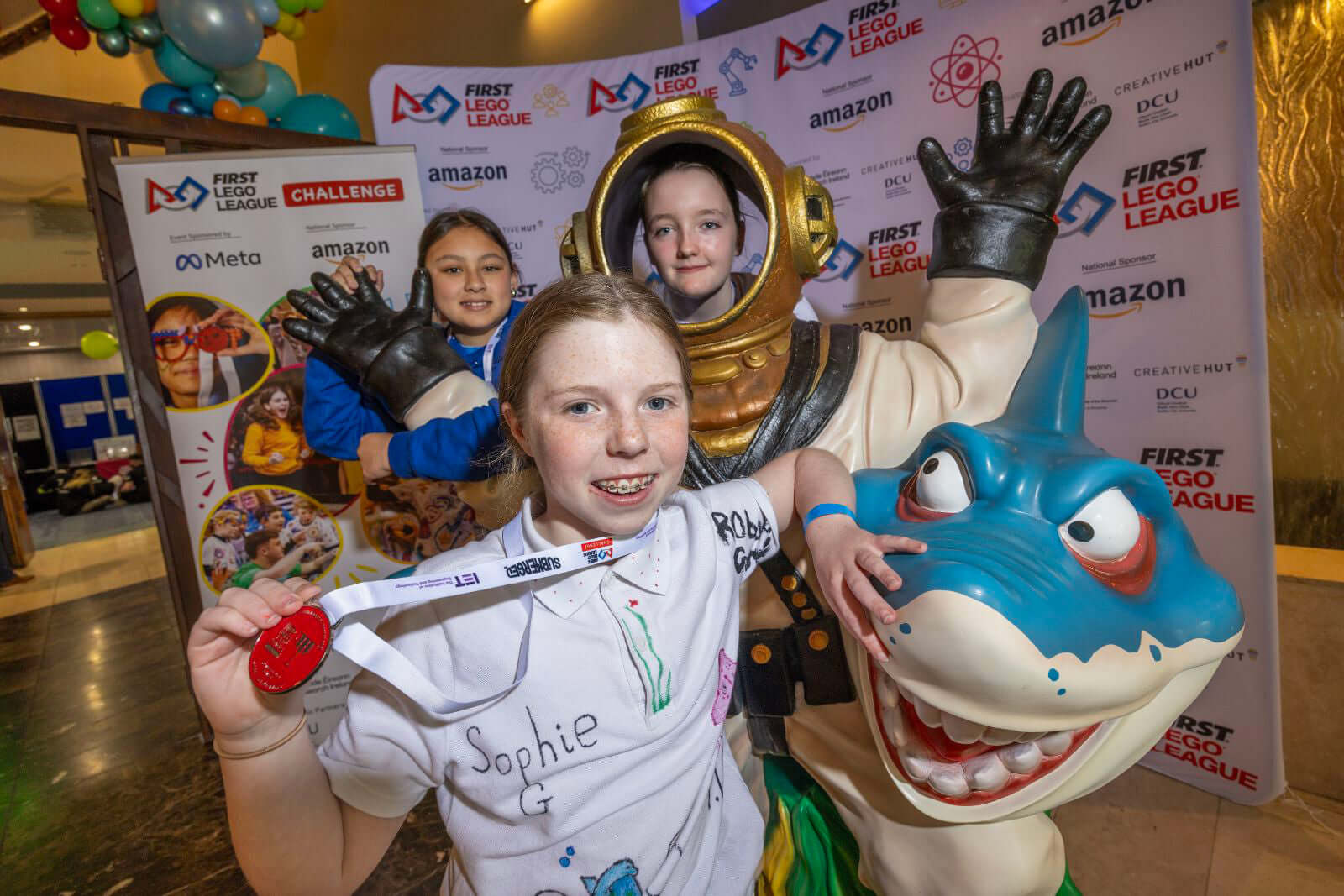 Team celebrating at the FIRST LEGO League event, featuring a girl with a medal, a diver mascot, and a shark costume.