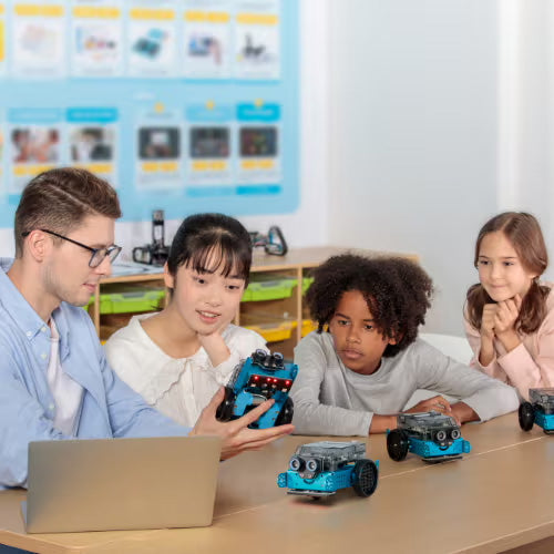 Students learning robotics with a teacher, showcasing robot models and a laptop in a classroom setting.