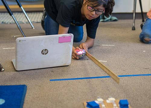 Student programming Sphero robot with laptop, ruler, and classroom materials during computer science lesson.