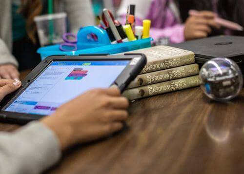 Student using a tablet to program concepts like loops and functions, with robotics books and tools on the table.