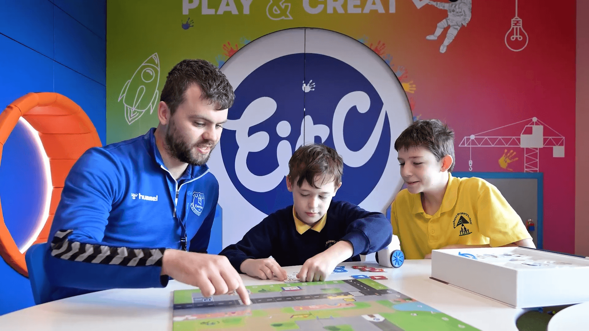 A teacher engages two children in a creative learning activity at a colorful classroom setting.