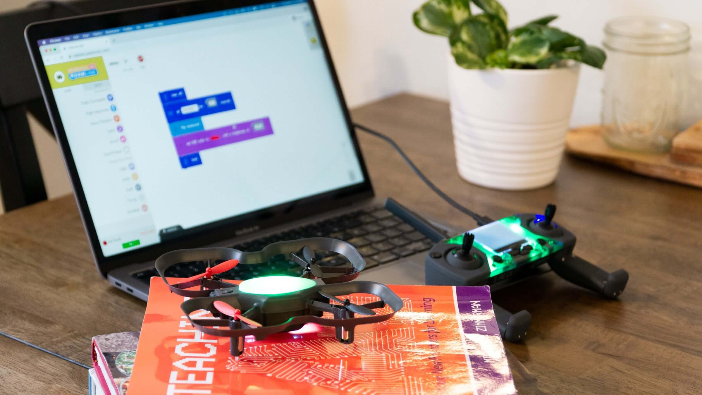 A laptop displays coding software next to a colorful drone and educational books on a wooden table.
