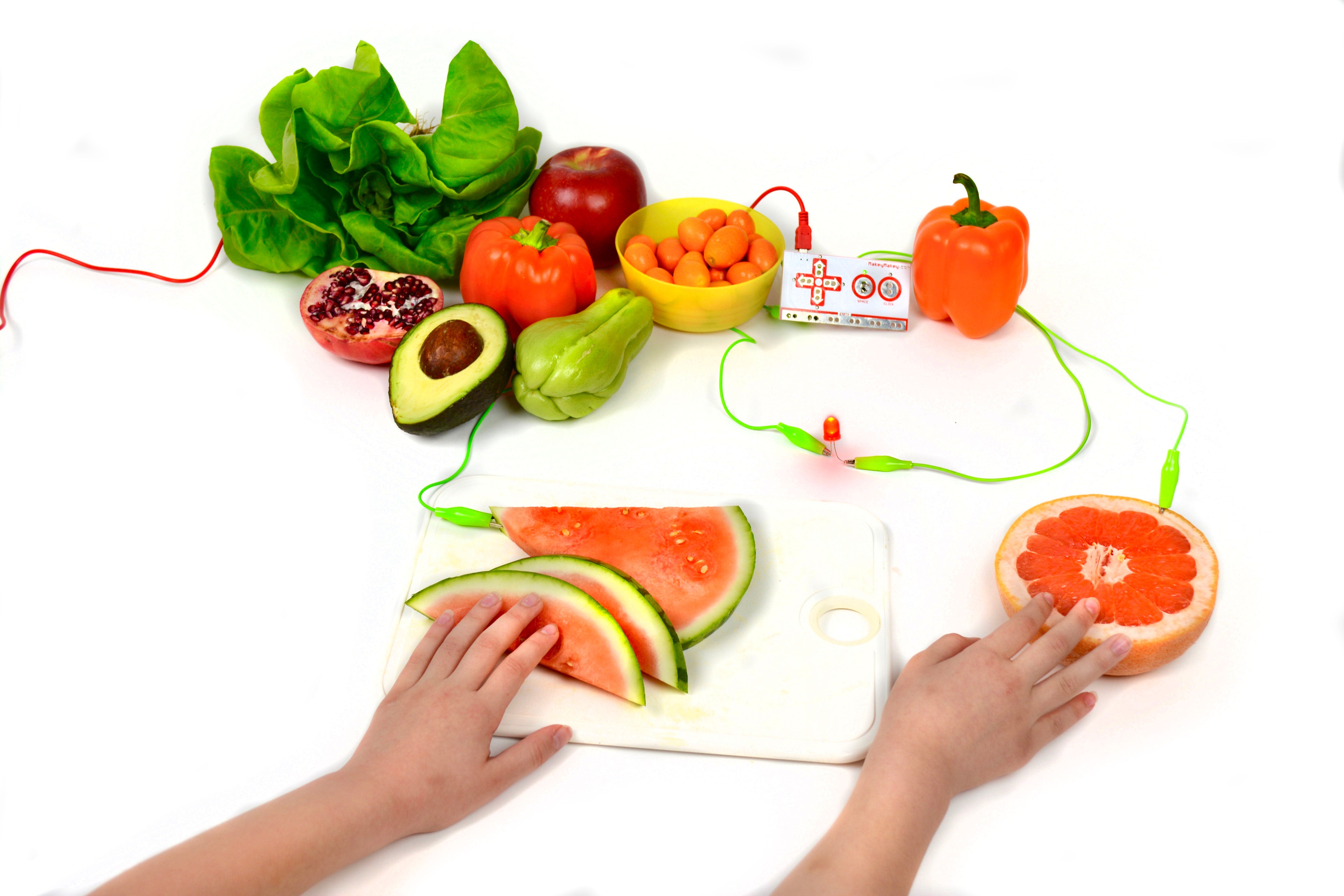 Colorful toy fruits and vegetables on a cutting board with a child's hands and battery connections for educational play.