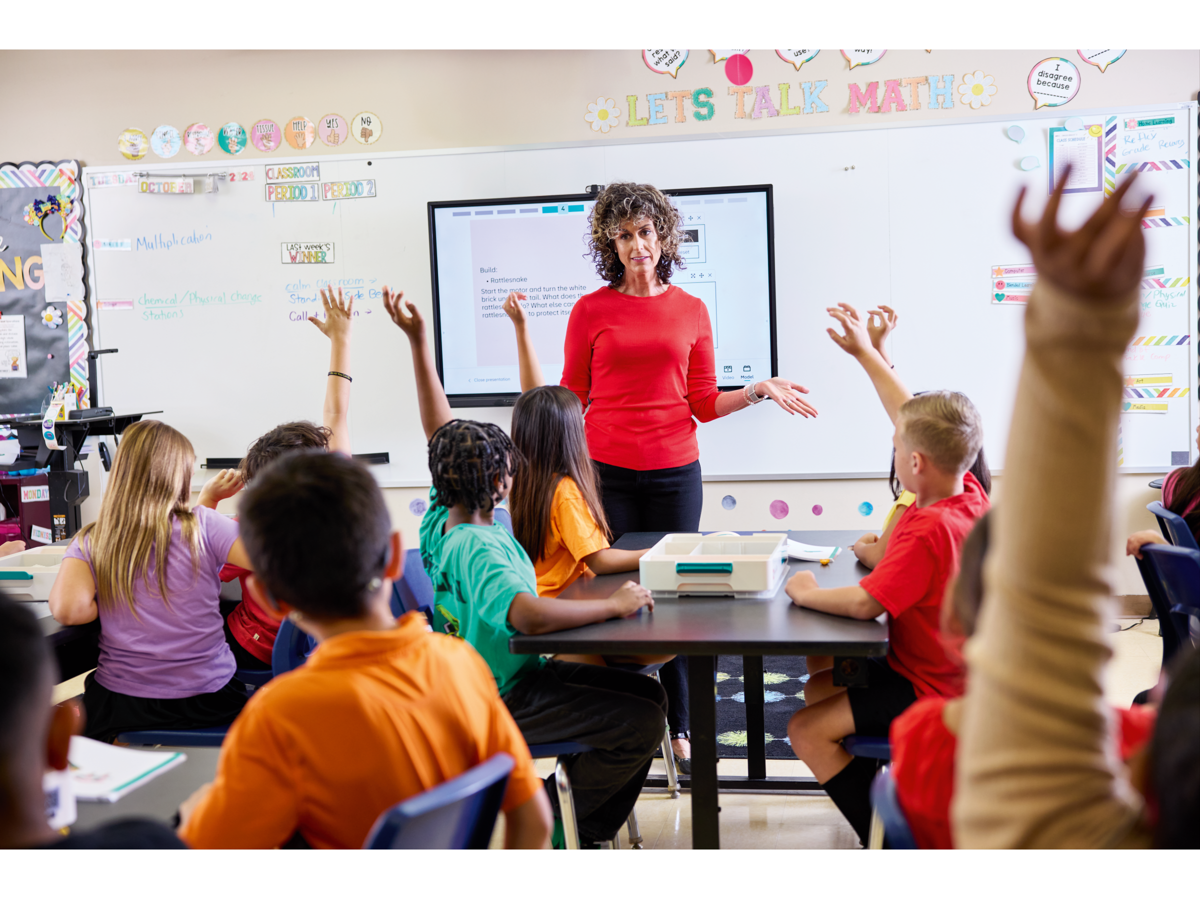 Engaged students raise hands in a lively classroom as their teacher explains math concepts on a smart board.