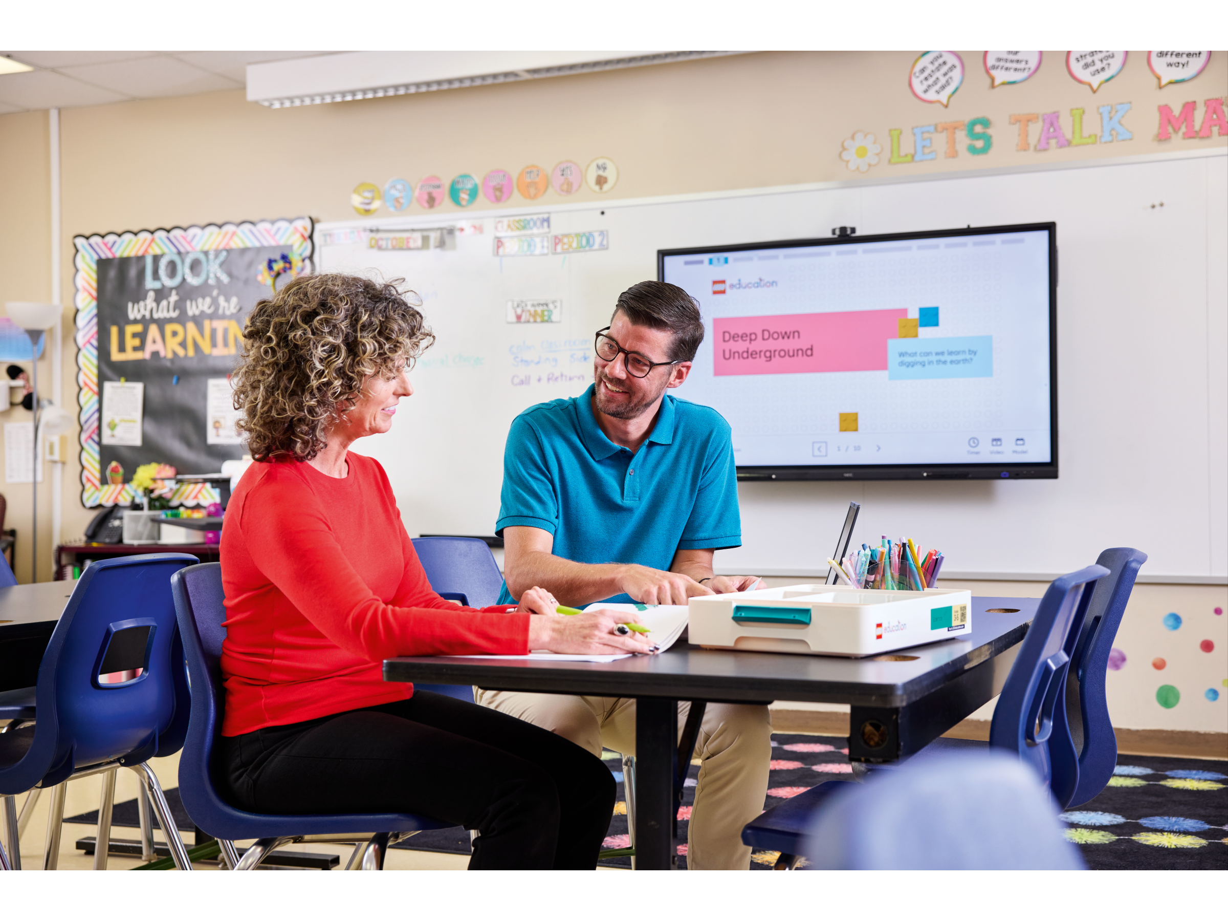 Two educators collaborating at a classroom table with a digital screen displaying educational content.
