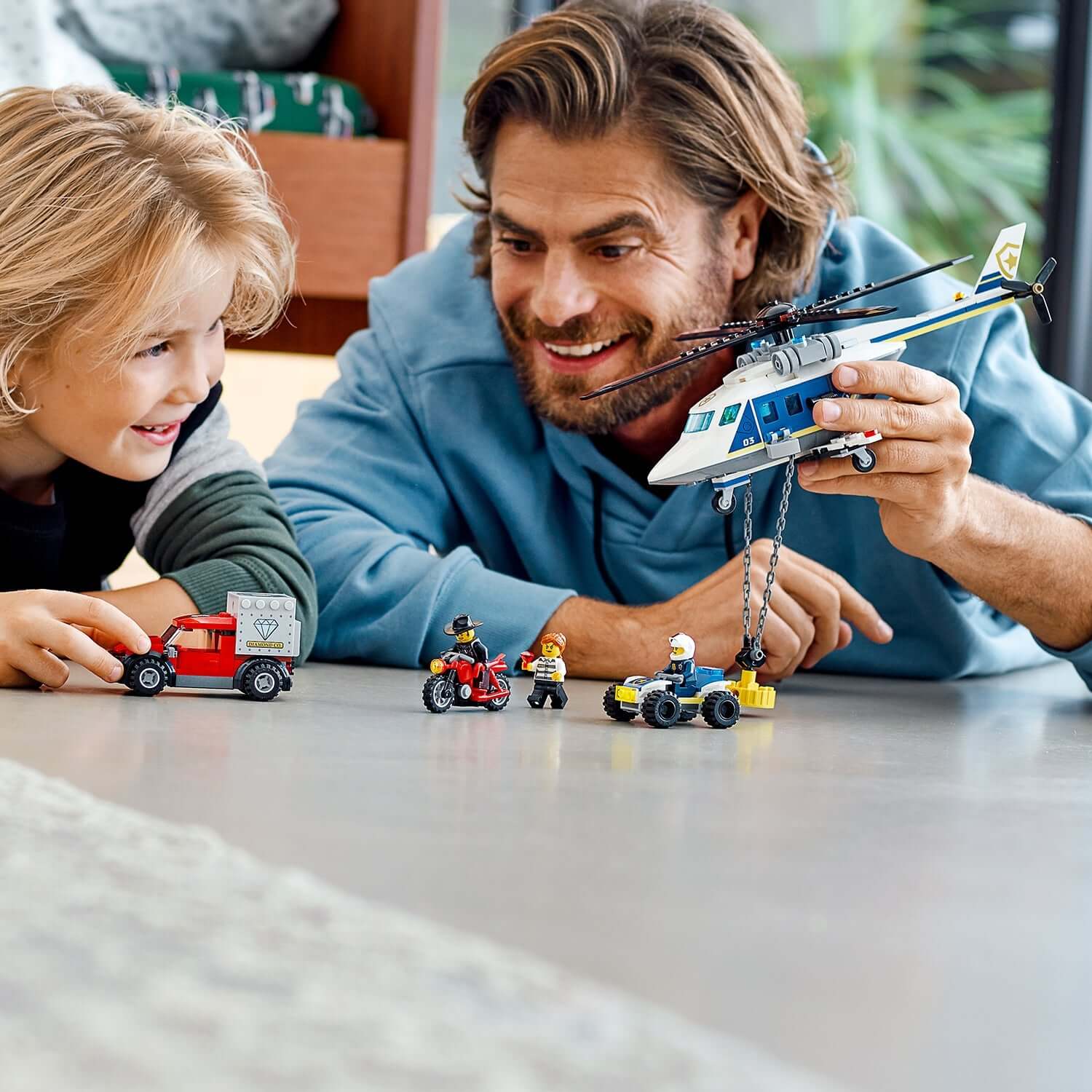 Child and adult playing with LEGO® City Police Helicopter Chase set, featuring helicopter and toy vehicles.