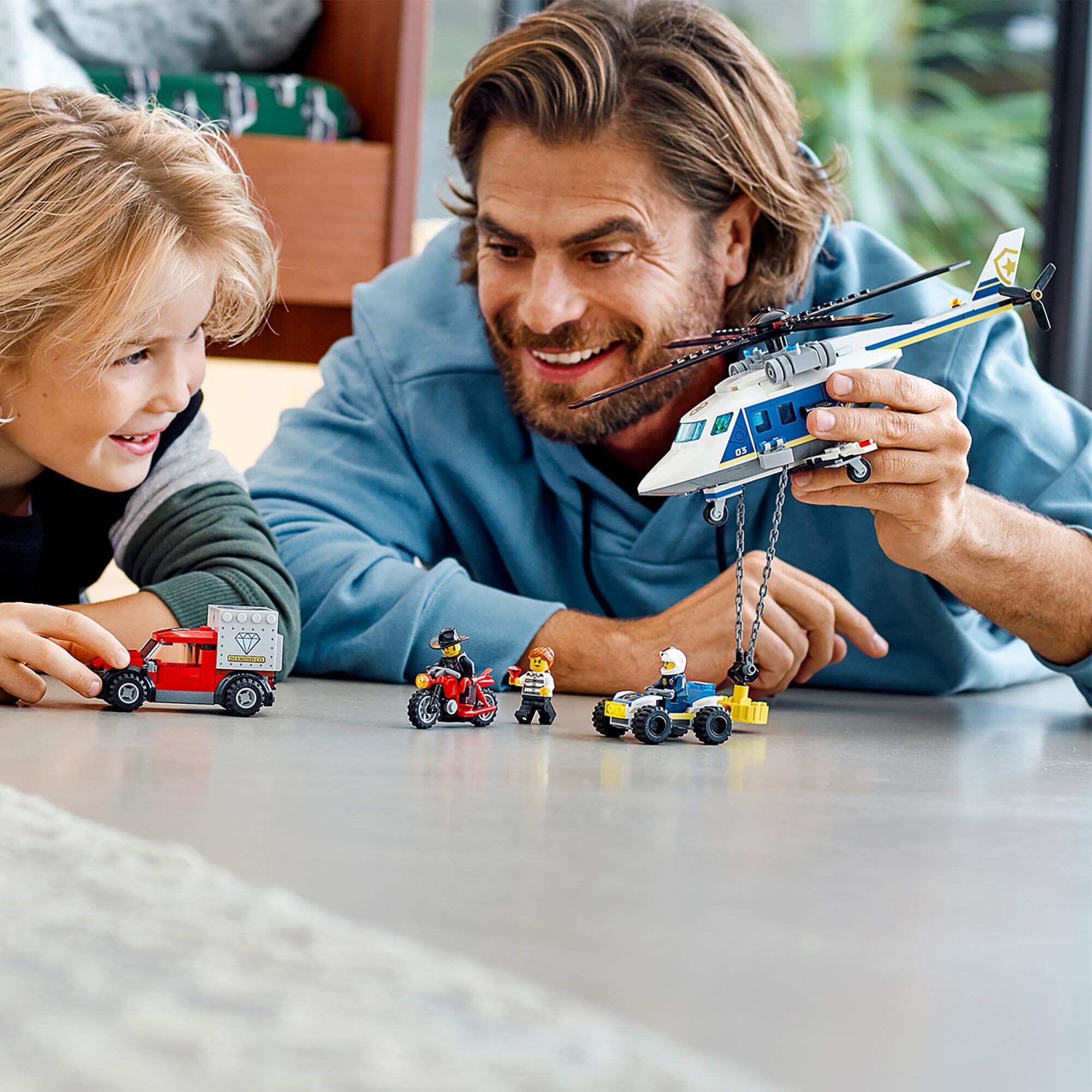 Father and son playing with LEGO® City Police Helicopter Chase set, featuring a helicopter and vehicles on a tabletop.