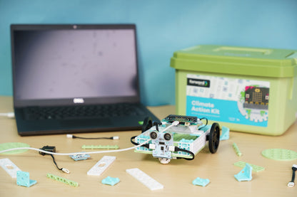 A table featuring a built robot vehicle from the Climate Action Kit, connected to a laptop. Scattered building components and a green storage bin labelled "Climate Action Kit" are visible on the wooden surface.
