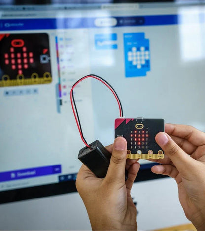 A close-up of a student’s hands holding a micro:bit V2 connected to a battery pack. The device’s LED matrix displays a red heart, mirroring the block-based coding interface visible on the laptop screen in the background.