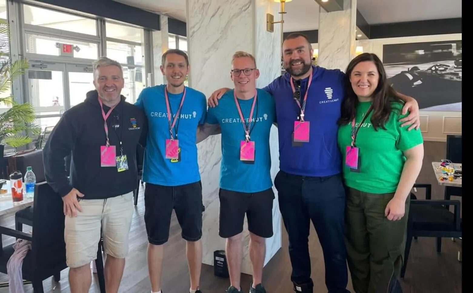Group of five smiling individuals in casual attire at an event, wearing lanyards and colorful shirts in a modern venue.