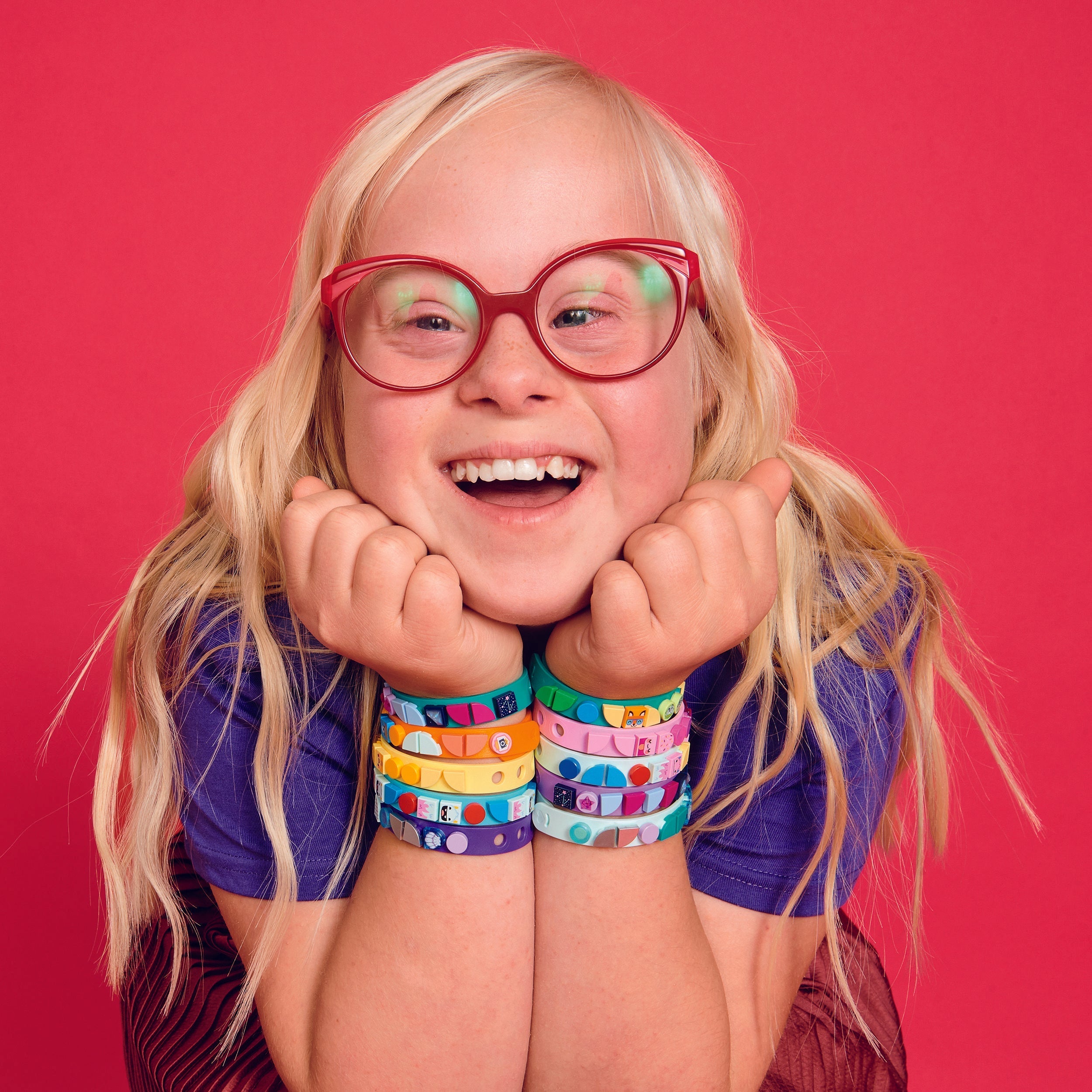 Child smiling with colorful LEGO® DOTS starlight bracelets on wrists against a vibrant pink background.
