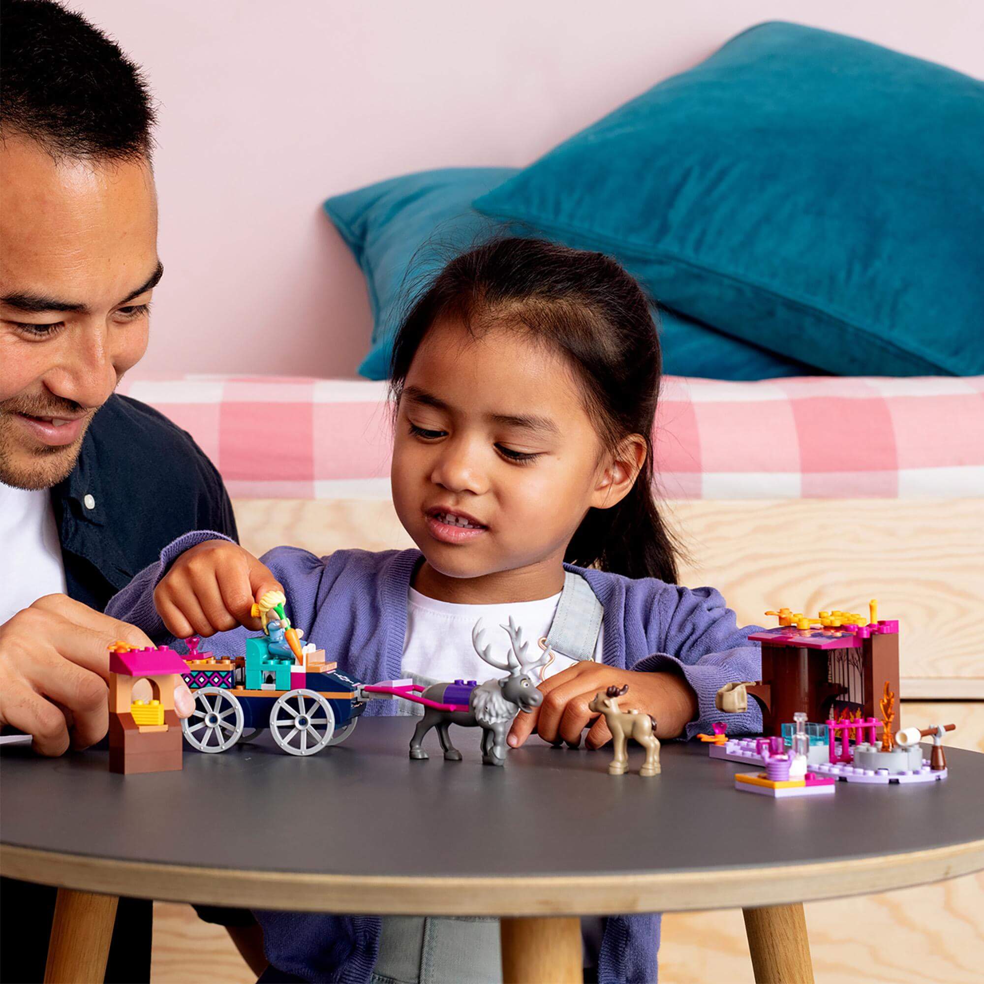 Child and parent playing with LEGO Disney Frozen 2 Elsa's Wagon Adventure set, featuring Elsa, her wagon, and animals.