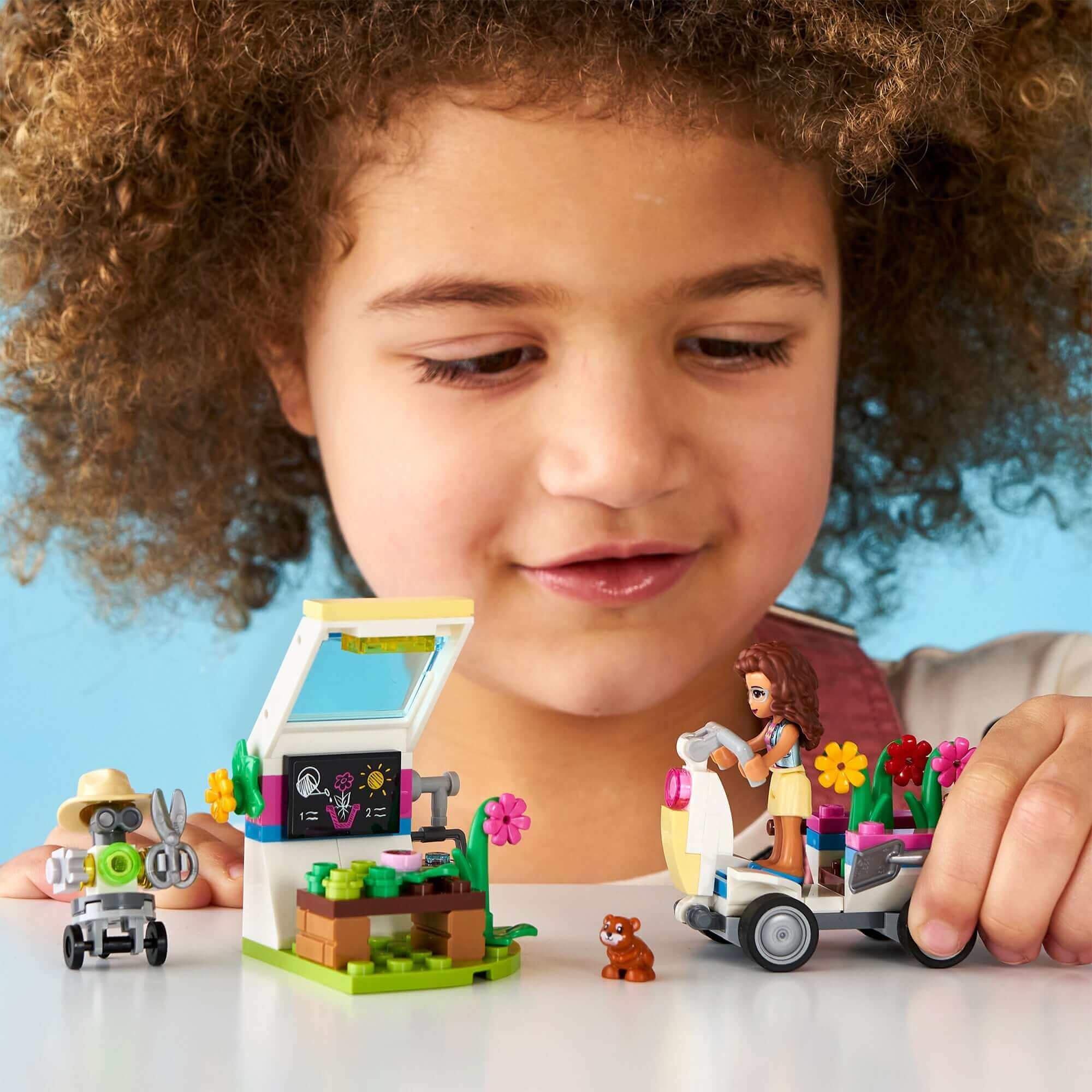 Child playing with LEGO Friends Olivia's Flower Garden set, featuring greenhouse, vehicle, and plant accessories.