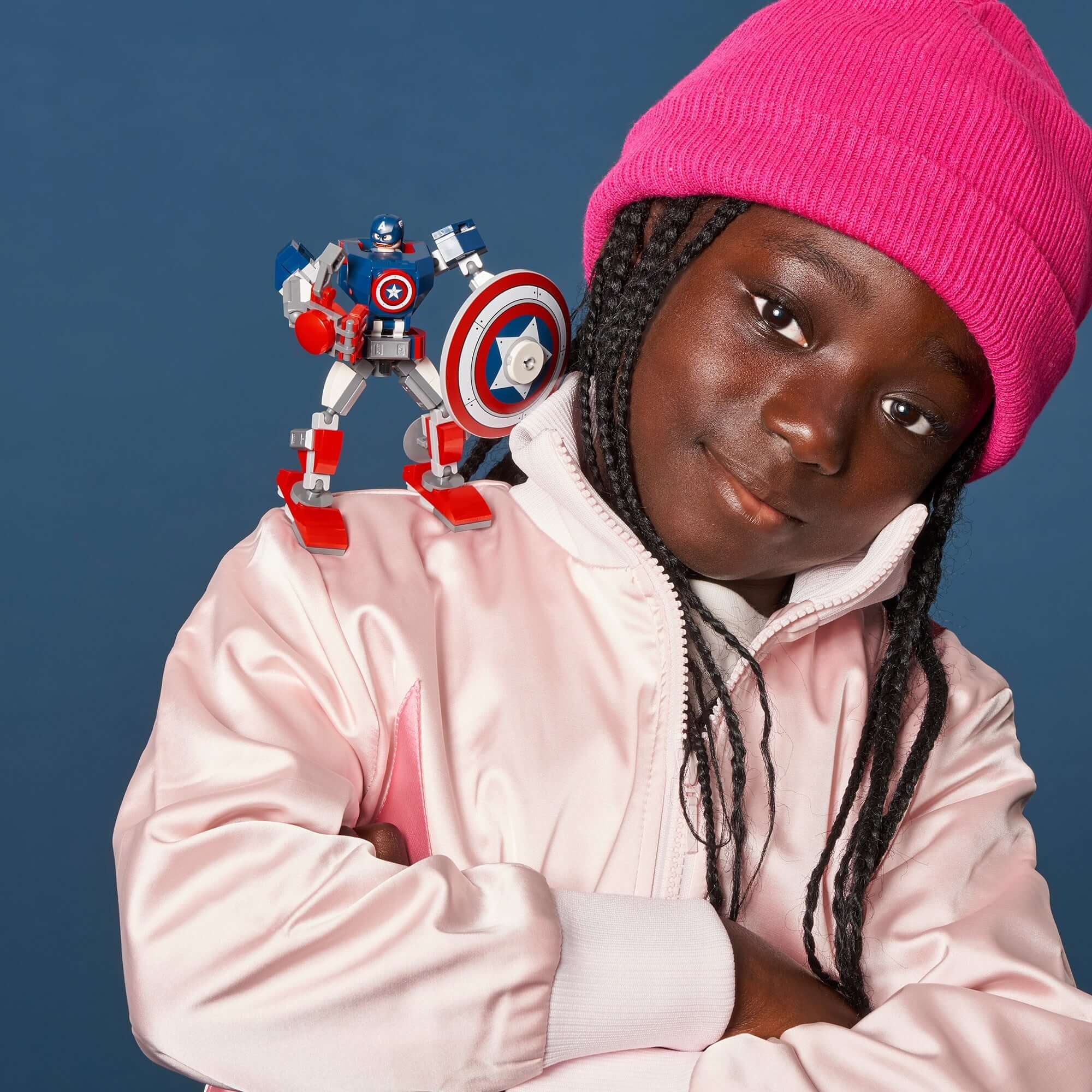 Child wearing a pink hat and jacket with a LEGO® Marvel Captain America mech on their shoulder, smiling for the camera.