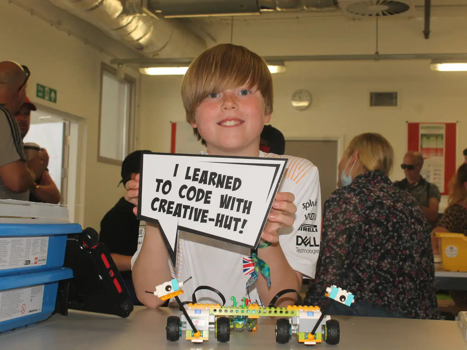 Smiling boy holding a sign saying 'I learned to code with Creative-Hut' next to a robotic creation.