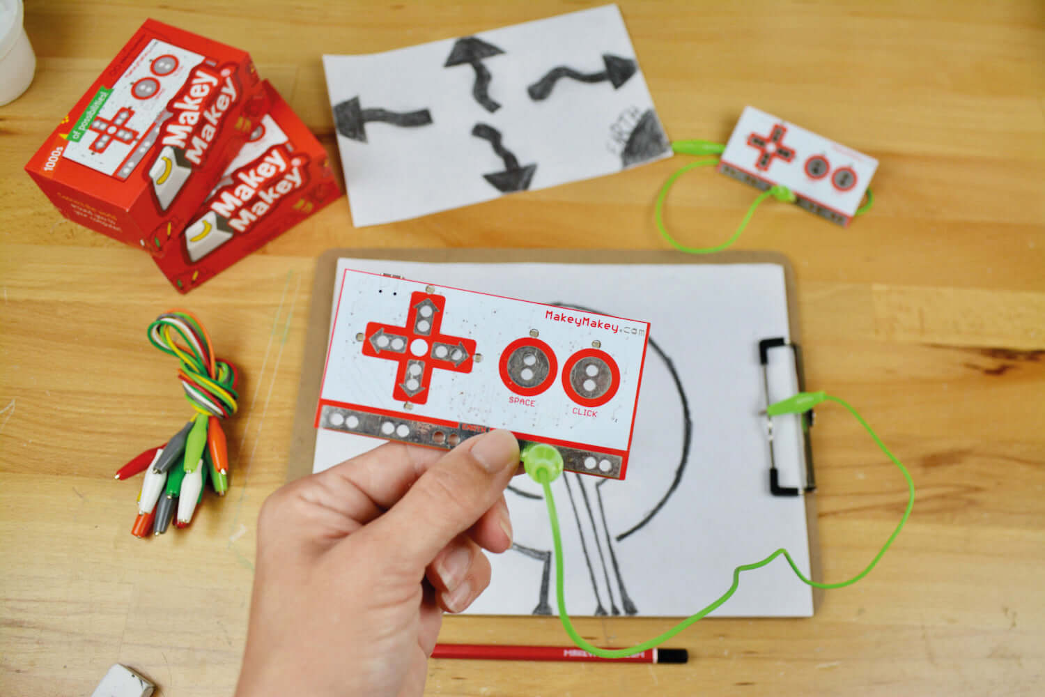 Person holding a Makey Makey kit with drawings and wires on a wooden table, showcasing interactive creativity.