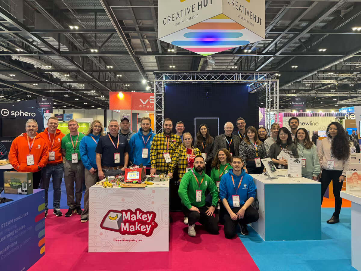 Group photo at Creative Hut booth showcasing Makey Makey at an event, featuring diverse participants and vibrant background.