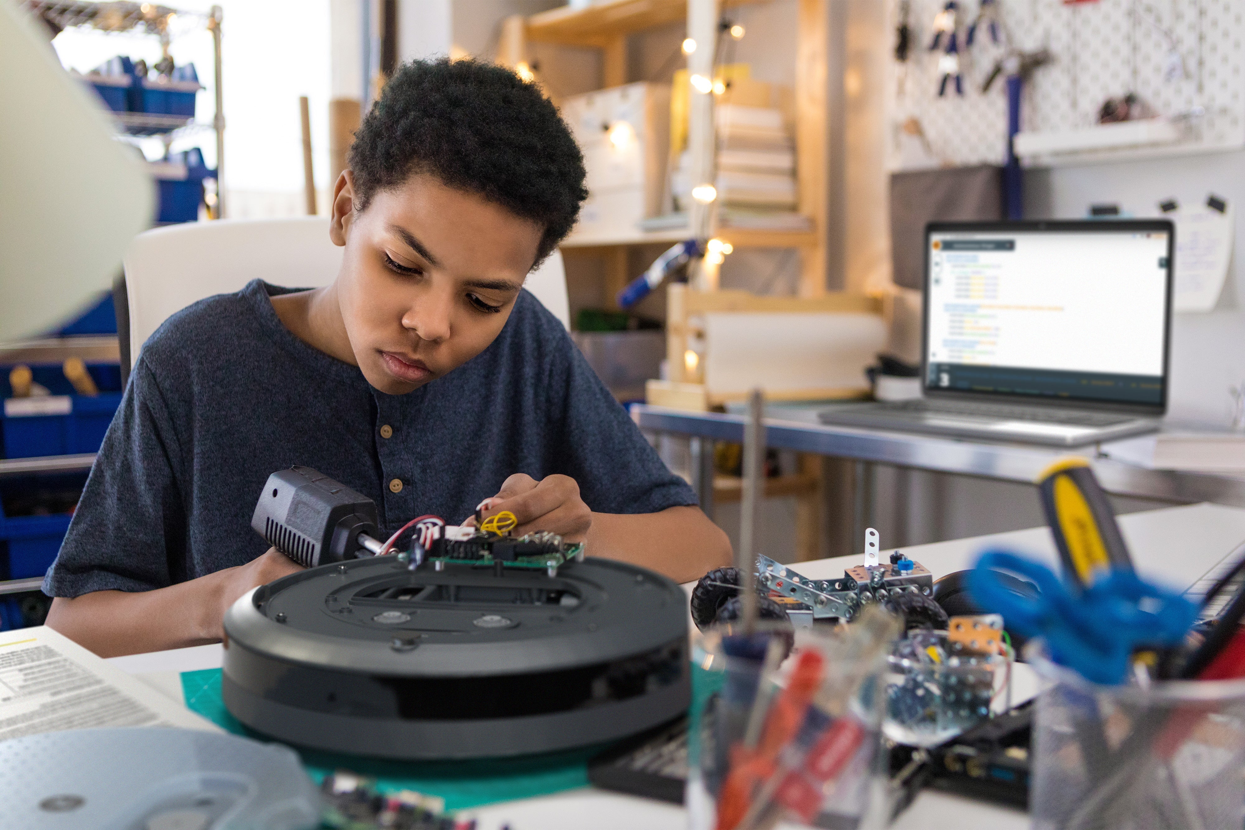 Young student programming the Create 3 educational robot in a creative workspace with tools and computer.