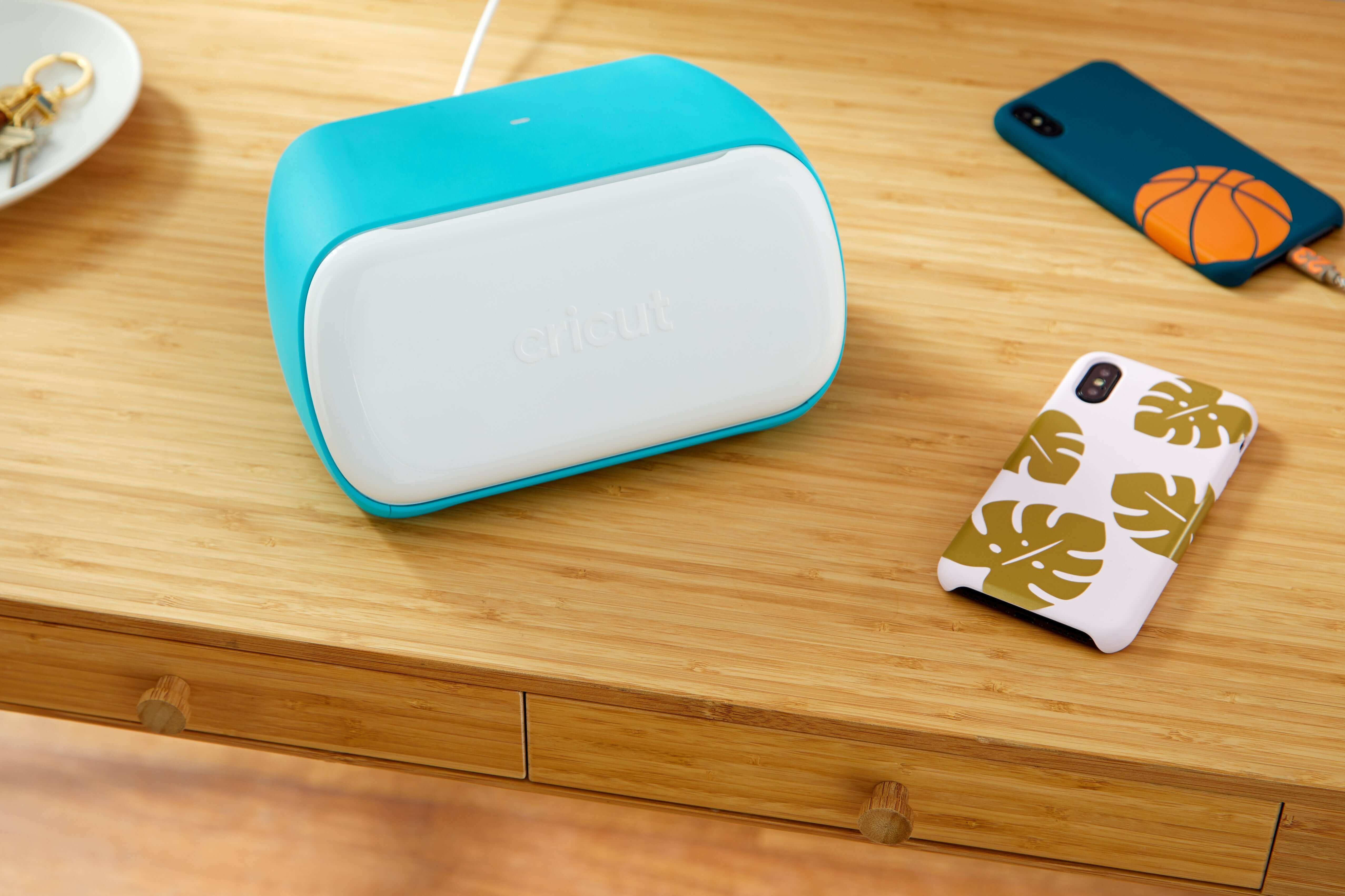 Cricut Joy cutting machine in teal color on a wooden desk with a personalized phone case.