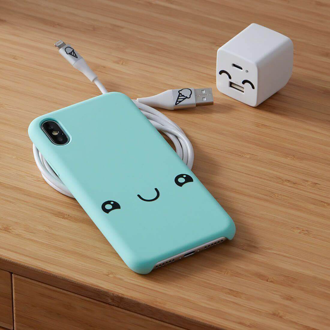 Cute mint phone case with a smiling face, charging cable, and a happy USB wall charger on a wooden surface.