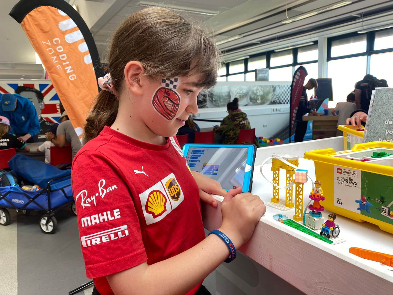 Child with face paint engaged in coding activity at a hands-on tech event, surrounded by colorful toys.