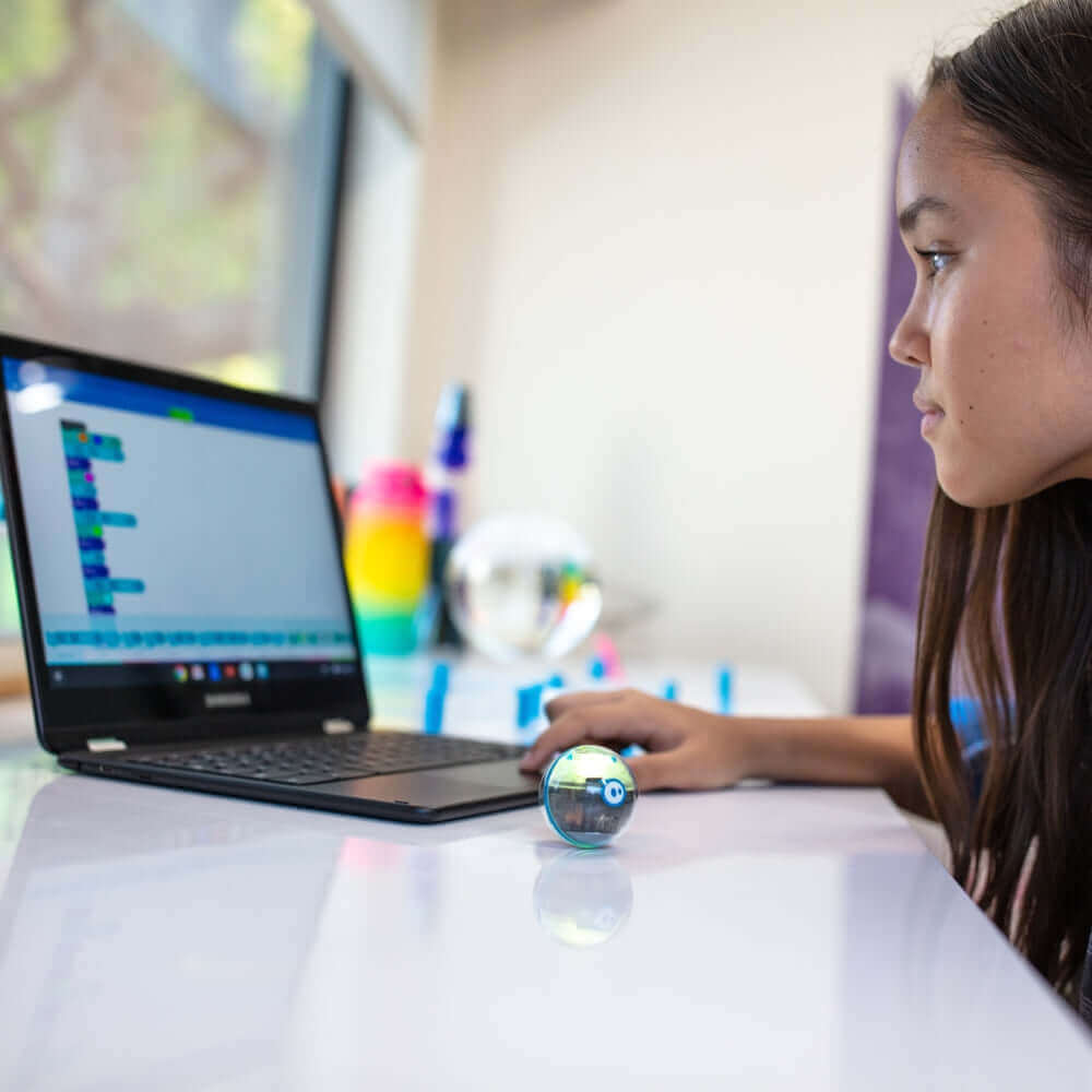 Young girl coding with Sphero Mini robot beside her on a desk, engaging with educational technology.