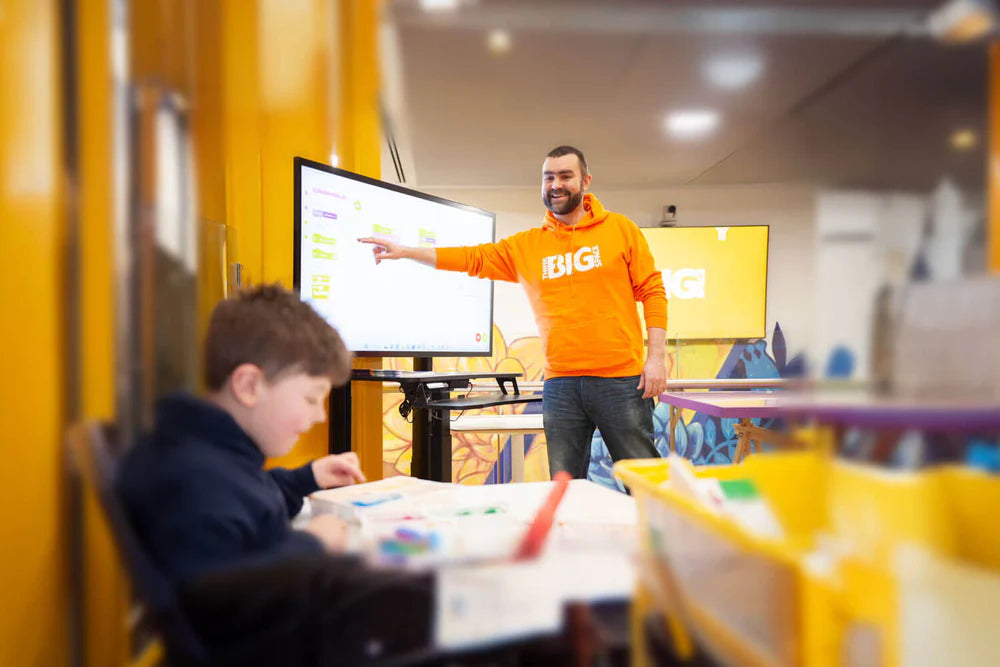 A teacher in an orange shirt engaging students with a presentation while a child works on art projects in a classroom.